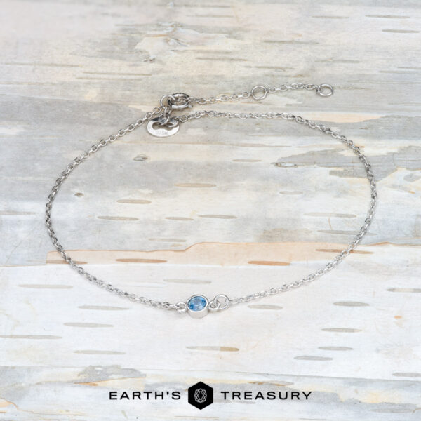 The “Lumi” Montana Sapphire Bracelet in 14k white gold A delicate bracelet crafted from 14k white gold, featuring a luminous Montana sapphire centerpiece, elegantly displayed on a light wooden surface. The bottom proudly showcases the brand name "Earth's Treasury.
