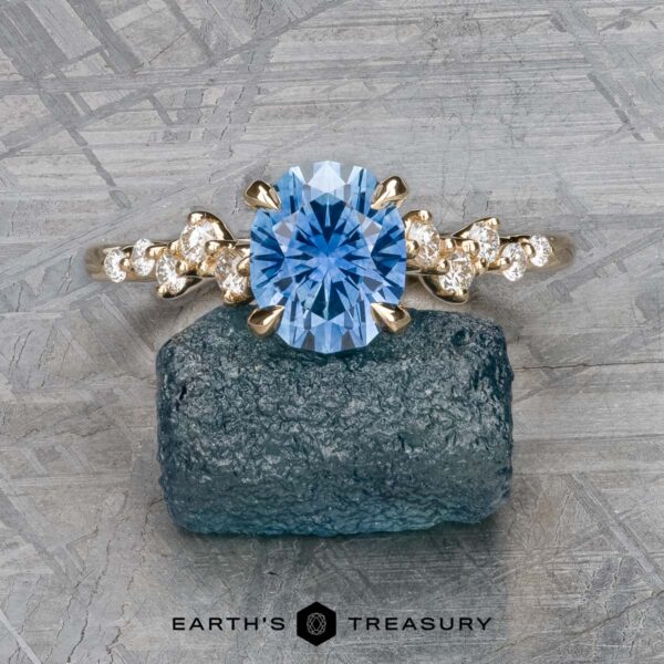 The "Catarina" ring in 14k yellow gold with 1.64-carat Montana sapphire The "Catarina" ring in 14k yellow gold, featuring a striking 1.64-carat Montana sapphire centerpiece surrounded by smaller clear stones, is showcased on a textured background in Primavera's intricate signature design.