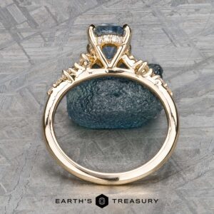 Set against a textured gray background, The "Catarina" ring in 14k yellow gold showcases a 1.64-carat Montana sapphire, elegantly surrounded by small radiant diamonds.