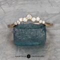 A stunning "Catarina" band in 14k yellow gold, featuring a hidden halo of small diamonds arranged in a chevron pattern, is elegantly displayed atop a blue gemstone. The backdrop showcases a textured gray surface with "Earth's Treasury" inscribed below, enhancing the sophisticated ambiance.
