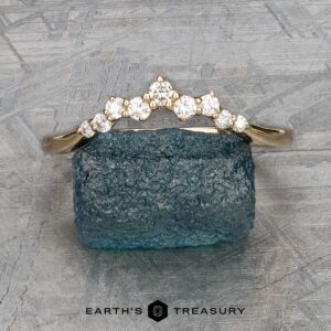 A stunning "Catarina" band in 14k yellow gold, featuring a hidden halo of small diamonds arranged in a chevron pattern, is elegantly displayed atop a blue gemstone. The backdrop showcases a textured gray surface with "Earth's Treasury" inscribed below, enhancing the sophisticated ambiance.