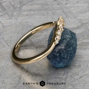 The "Catarina" band in 14k yellow gold, adorned with diamonds, elegantly rests against a rough blue stone on a textured gray surface.