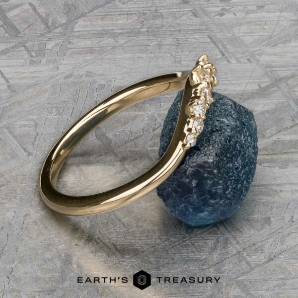 The "Catarina" band in 14k yellow gold The "Catarina" band in 14k yellow gold, adorned with diamonds, elegantly rests against a rough blue stone on a textured gray surface.