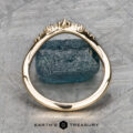 The "Catarina" band in 14k yellow gold, featuring a jagged, textured design that captures Catarina's rugged beauty, is elegantly displayed upright before a rough blue gemstone. The backdrop showcases a metallic cross-hatched pattern, and the "Earth's Treasury" logo shimmers beneath like the subtle charm of a hidden halo ring.