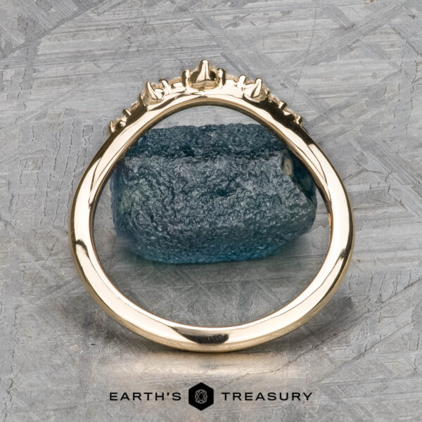 The "Catarina" band in 14k yellow gold The "Catarina" band in 14k yellow gold, featuring a jagged, textured design that captures Catarina's rugged beauty, is elegantly displayed upright before a rough blue gemstone. The backdrop showcases a metallic cross-hatched pattern, and the "Earth's Treasury" logo shimmers beneath like the subtle charm of a hidden halo ring.
