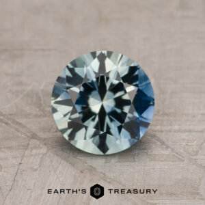 1.23-carat aqua-blue particolored Montana Sapphire (heated) with blue and green hues shown on a textured surface, "EARTH'S TREASURY" logo and text below.