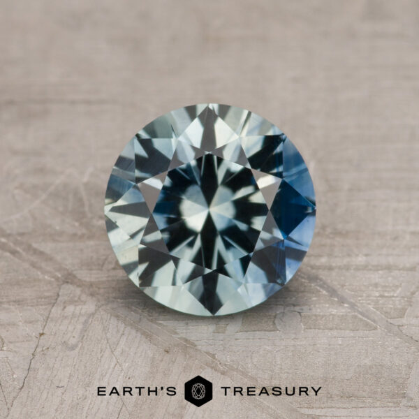 1.23-Carat Aqua-Blue Particolored Montana Sapphire (Heated) 1.23-carat aqua-blue particolored Montana Sapphire (heated) with blue and green hues shown on a textured surface, "EARTH'S TREASURY" logo and text below.