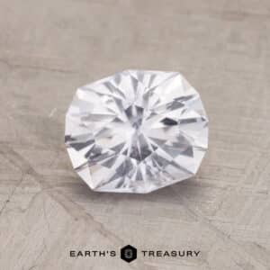 A 1.75-carat near colorless Montana sapphire with a brilliant cut is displayed against a light grey textured background. The gemstone's intricate facets reflect light, creating a sparkling effect. Below the gemstone, the text "EARTH'S TREASURY" appears in black, along with a small geometric icon.