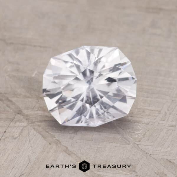 1.75-Carat Montana Sapphire (Heated) A 1.75-carat near colorless Montana sapphire with a brilliant cut is displayed against a light grey textured background. The gemstone's intricate facets reflect light, creating a sparkling effect. Below the gemstone, the text "EARTH'S TREASURY" appears in black, along with a small geometric icon.
