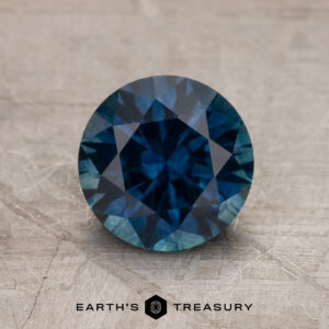 A 1.77-carat Deep Blue Montana Sapphire (heated) with a round brilliant cut is displayed on a textured surface, featuring the text "EARTH'S TREASURY" below it.
