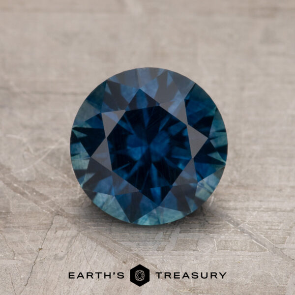 1.77-Carat Deep Blue Montana Sapphire (Heated) A 1.77-carat Deep Blue Montana Sapphire (heated) with a round brilliant cut is displayed on a textured surface, featuring the text "EARTH'S TREASURY" below it.