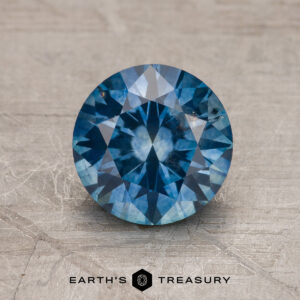 A 1.75-carat heated Montana Sapphire in medium blue, cut in a round brilliant style, is displayed on a textured surface with the "Earth's Treasury" logo beneath it.