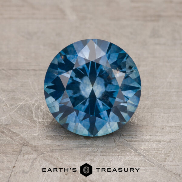 1.75-Carat Montana Sapphire (Heated) A 1.75-carat heated Montana Sapphire in medium blue, cut in a round brilliant style, is displayed on a textured surface with the "Earth's Treasury" logo beneath it.
