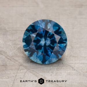 A 1.66-carat Peacock Blue Montana Sapphire (Heated), round brilliant-cut, rests on a textured gray surface featuring the Earth's Treasury logo and text below.