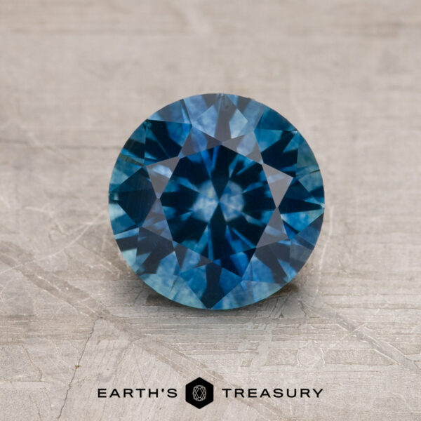 1.66-Carat Peacock Blue Montana Sapphire (Heated) A 1.66-carat Peacock Blue Montana Sapphire (Heated), round brilliant-cut, rests on a textured gray surface featuring the Earth's Treasury logo and text below.