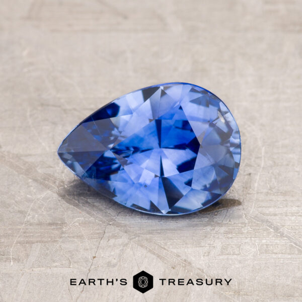 1.63-Carat Rich Cornflower Blue Ceylon Sapphire (Heated) A pear-shaped blue gemstone with multi-faceted cuts is displayed on a textured surface. The text "1.63-Carat Ceylon Sapphire (Heated)" is visible at the bottom of the image.