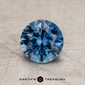 A 1.37-carat medium blue Montana Sapphire (heated), round brilliant-cut, is shown on a textured gray surface with "EARTH'S TREASURY" displayed below.