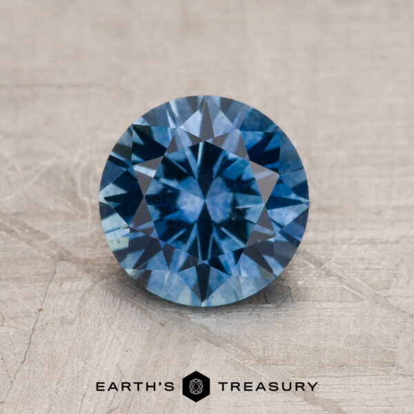 1.37-Carat Medium Blue Montana Sapphire (Heated) A 1.37-carat medium blue Montana Sapphire (heated), round brilliant-cut, is shown on a textured gray surface with "EARTH'S TREASURY" displayed below.