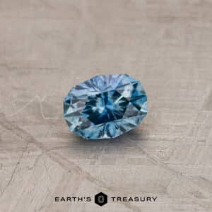 A 0.85-carat medium blue Montana sapphire, oval-shaped and faceted, rests on a textured gray surface. The gemstone showcases various shades of blue with intricate inner patterns. Below the stone, the text "Earth's Treasury" is visible.