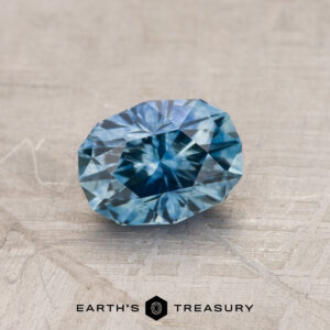 A 0.85-carat medium blue Montana sapphire, oval-shaped and faceted, rests on a textured gray surface. The gemstone showcases various shades of blue with intricate inner patterns. Below the stone, the text "Earth's Treasury" is visible.