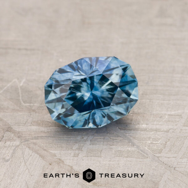 0.85-Carat Montana Sapphire (Heated) A 0.85-carat medium blue Montana sapphire, oval-shaped and faceted, rests on a textured gray surface. The gemstone showcases various shades of blue with intricate inner patterns. Below the stone, the text "Earth's Treasury" is visible.