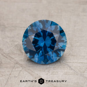 A 1.24-Carat Rich Blue Montana Sapphire (Heated), round and faceted, is shown on a textured gray surface with the Earth's Treasury logo at the bottom of the image.