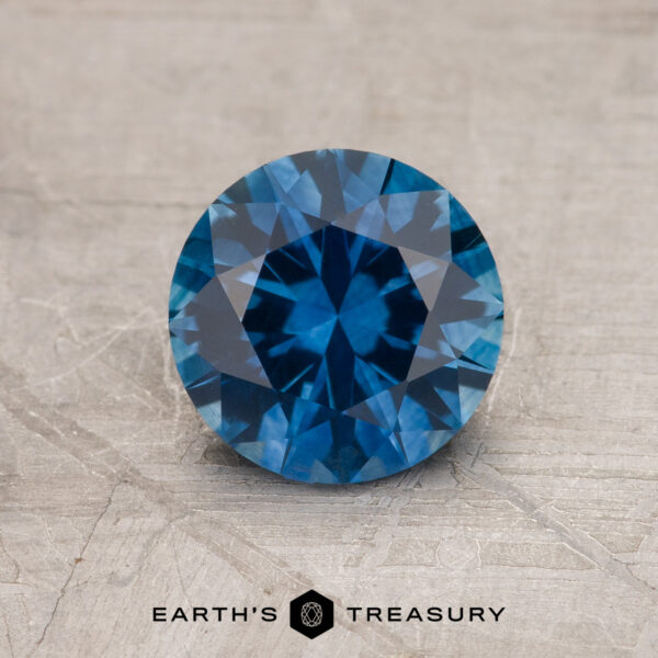 1.24-Carat Rich Blue Montana Sapphire (Heated) A 1.24-Carat Rich Blue Montana Sapphire (Heated), round and faceted, is shown on a textured gray surface with the Earth's Treasury logo at the bottom of the image.