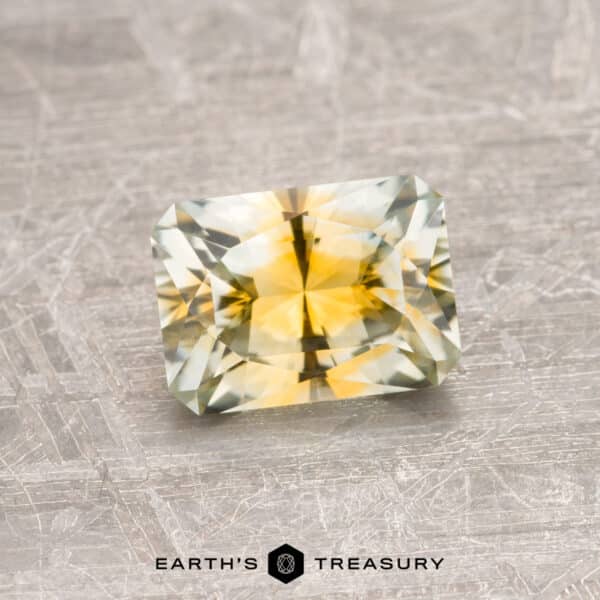 1.41-Carat Montana Sapphire (Heated) A 1.41-carat Yellow-Gold Particolored Montana Sapphire, featuring a radiant cut, rests on a textured surface. The gemstone displays an exquisite blend of yellow and green hues and is expertly faceted to catch the light. At the bottom of the image, the text "EARTH'S TREASURY" appears alongside a hexagonal logo.
