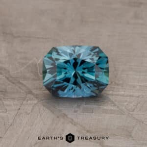 A multifaceted, rectangular, 1.12-carat rich teal Montana sapphire (heated) is displayed against a textured, light background. The text "EARTH'S TREASURY" appears beneath the gemstone in black.