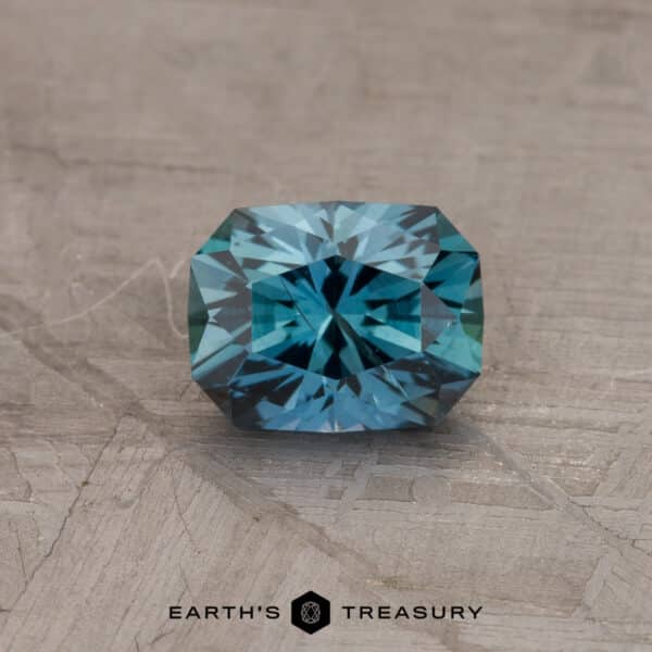 1.12-Carat Montana Sapphire (Heated) A multifaceted, rectangular, 1.12-carat rich teal Montana sapphire (heated) is displayed against a textured, light background. The text "EARTH'S TREASURY" appears beneath the gemstone in black.