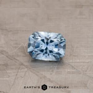 A radiant-cut Sky Blue Montana Sapphire (0.87 carats, heated) with intricate facets reflecting light, placed on a textured beige surface. The bottom of the image displays the text "EARTH'S TREASURY" alongside a hexagonal logo.