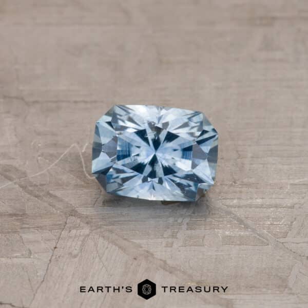 0.87-Carat Montana Sapphire (Heated) A radiant-cut Sky Blue Montana Sapphire (0.87 carats, heated) with intricate facets reflecting light, placed on a textured beige surface. The bottom of the image displays the text "EARTH'S TREASURY" alongside a hexagonal logo.