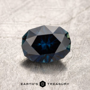 A 1.27-carat Australian Sapphire, faceted in Midnight Blue, rests on a textured, light-colored surface with the "EARTH'S TREASURY" logo and text beneath it.