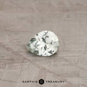 A faceted 0.73-carat pale mint green Montana sapphire, shaped like a pear, rests on a textured surface. The light-colored gem appears almost translucent with various facets catching the light. Below the sapphire, the text reads "EARTH'S TREASURY" accompanied by a hexagonal logo.