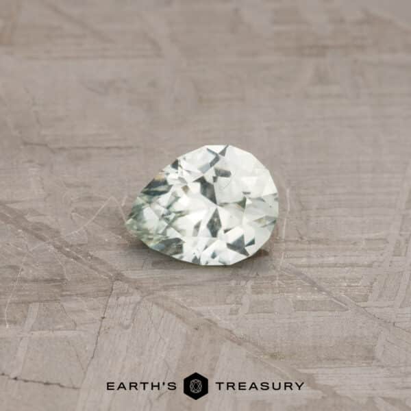 0.73-Carat Montana Sapphire (Heated) A faceted 0.73-carat pale mint green Montana sapphire, shaped like a pear, rests on a textured surface. The light-colored gem appears almost translucent with various facets catching the light. Below the sapphire, the text reads "EARTH'S TREASURY" accompanied by a hexagonal logo.