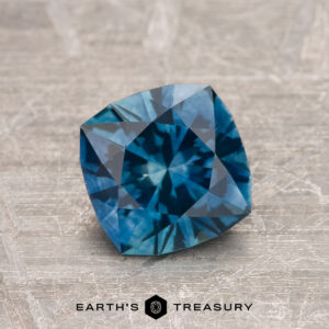 A 1.34-carat Peacock Blue Montana Sapphire (Heated) with square cushion cut and intricate facets is displayed on a textured surface, with the Earth’s Treasury logo visible at the bottom.