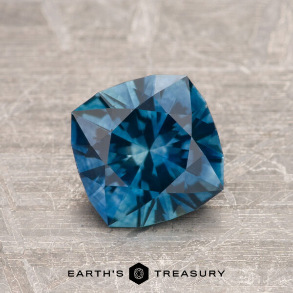 1.34-Carat Peacock Blue Montana Sapphire (Heated) A 1.34-carat Peacock Blue Montana Sapphire (Heated) with square cushion cut and intricate facets is displayed on a textured surface, with the Earth’s Treasury logo visible at the bottom.