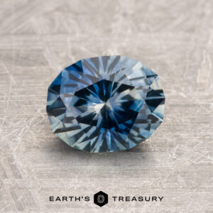 A 1.80-carat blue-aqua particolored Montana Sapphire (heated), round and faceted, displayed on a textured surface with "EARTH'S TREASURY" below.