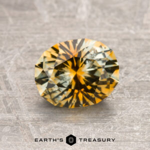 A 1.75-carat yellow-orange particolored Montana Sapphire (heated) is displayed on a textured surface with the "Earth's Treasury" logo and text below it.
