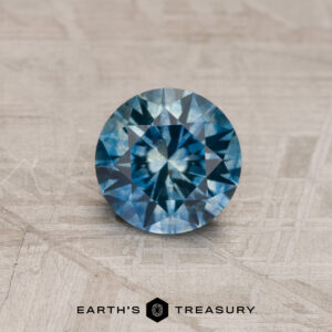A 1.54-carat Teal Blue Montana Sapphire (Heated), cut in a round brilliant shape, rests on a textured surface with "Earth's Treasury" beneath it.