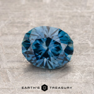 A 1.41-carat teal blue Montana sapphire (heated) with striking facets sits on a textured metallic surface, with the Earth's Treasury logo below.