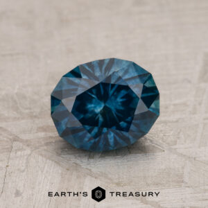 A 1.46-carat deep blue-green Montana Sapphire (heated), oval-cut, rests on a textured surface with the "EARTH'S TREASURY" logo visible at the bottom of the image.