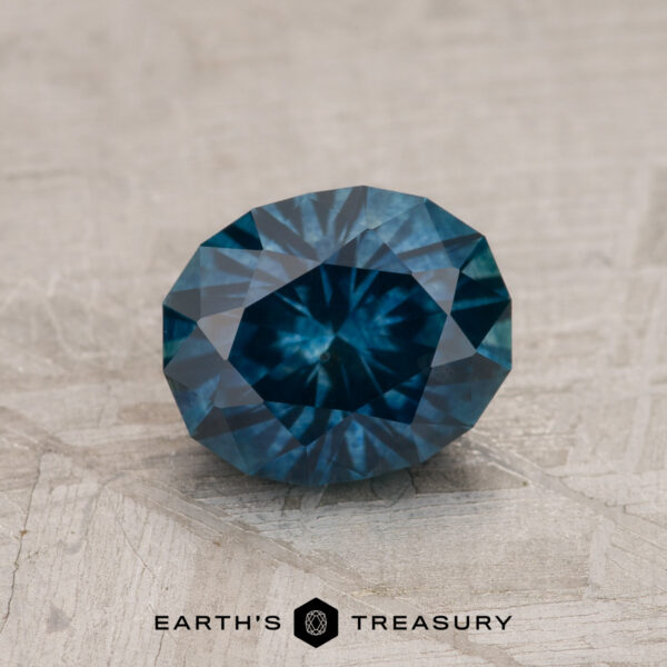 1.46-Carat Deep Blue-Green Montana Sapphire (Heated) A 1.46-carat deep blue-green Montana Sapphire (heated), oval-cut, rests on a textured surface with the "EARTH'S TREASURY" logo visible at the bottom of the image.