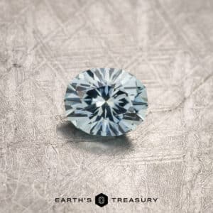 A sparkling round-cut 1.20-carat light aqua blue Montana sapphire with precise facets sits on a textured gray surface. Below the gemstone, "EARTH'S TREASURY" is written in black text, accompanied by a small geometric logo.