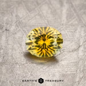 A 1.27-carat, yellow-gold particolored Montana sapphire (heated) sits on a textured metallic surface. Below the gemstone, the text "EARTH'S TREASURY" is displayed alongside a small hexagonal logo. The sapphire boasts bright, reflective facets and a rich, vibrant yellow color.