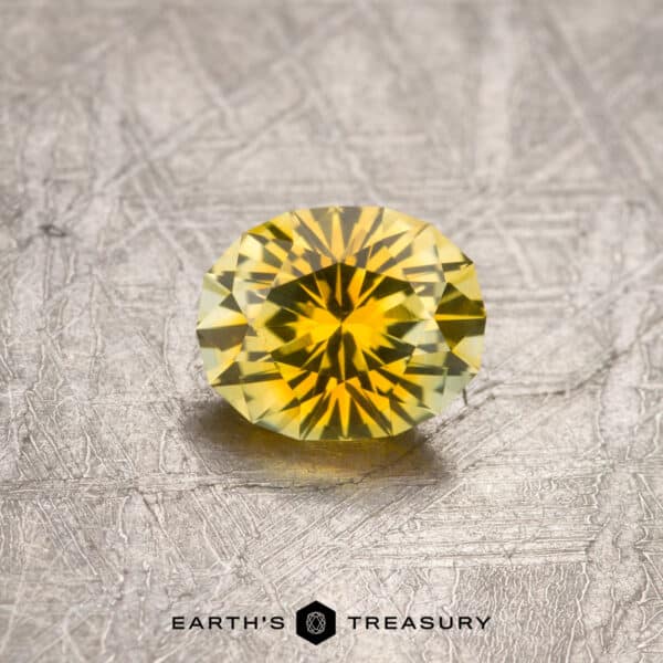 1.27-Carat Montana Sapphire (Heated) A 1.27-carat, yellow-gold particolored Montana sapphire (heated) sits on a textured metallic surface. Below the gemstone, the text "EARTH'S TREASURY" is displayed alongside a small hexagonal logo. The sapphire boasts bright, reflective facets and a rich, vibrant yellow color.