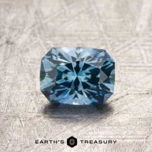 A 1.02-carat rich Sky Blue Montana Sapphire (heated), faceted in a rectangular shape with a starburst pattern, is displayed on a textured surface above the Earth’s Treasury logo.