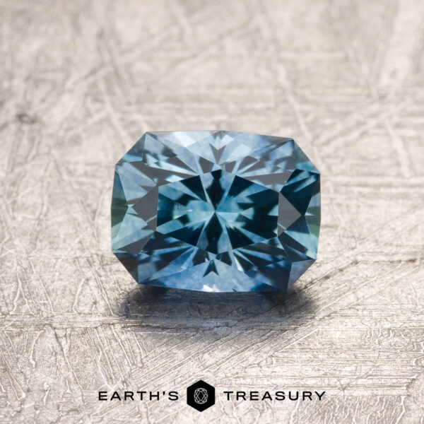 1.02-Carat Rich Sky Blue Montana Sapphire (Heated) A 1.02-carat rich Sky Blue Montana Sapphire (heated), faceted in a rectangular shape with a starburst pattern, is displayed on a textured surface above the Earth’s Treasury logo.