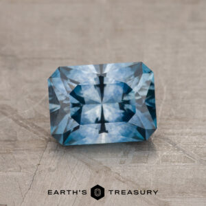 A 1.42-carat medium blue Montana Sapphire (heated), rectangular in shape, is shown on a textured gray surface with "EARTH'S TREASURY" written below it.
