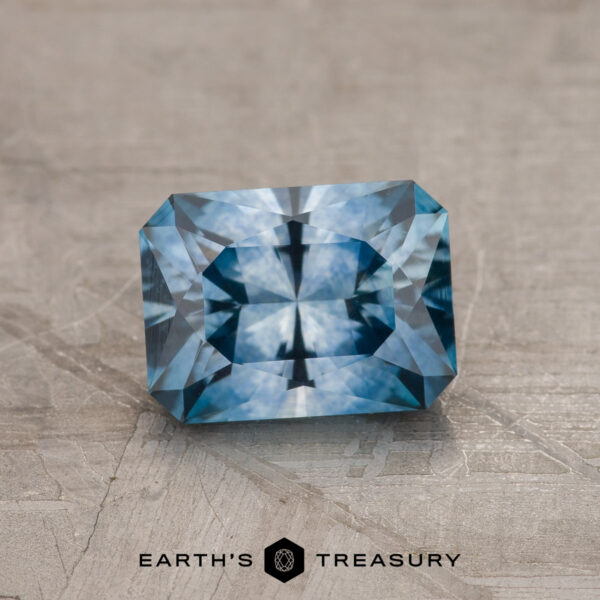 1.42-Carat Medium Blue Montana Sapphire (Heated) A 1.42-carat medium blue Montana Sapphire (heated), rectangular in shape, is shown on a textured gray surface with "EARTH'S TREASURY" written below it.